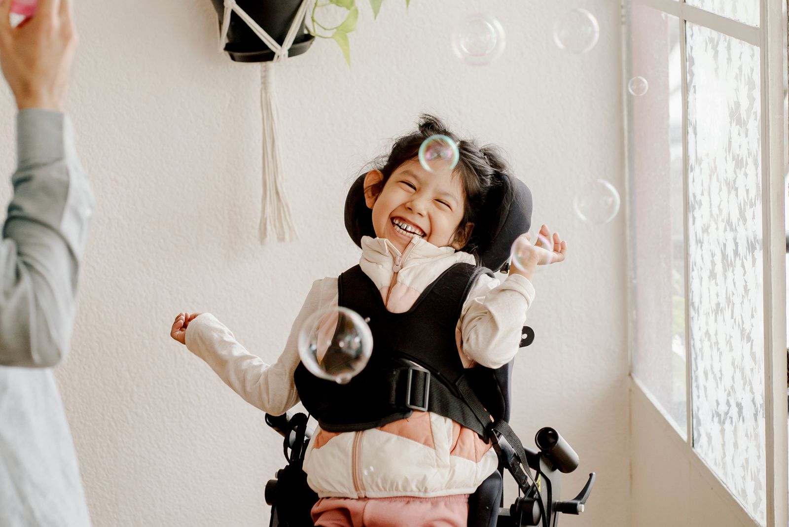 A smiling child in an adaptive chair laughing as bubbles float around her